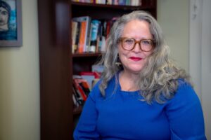 M. Soledad Caballero is facing the viewer, smiling, wearing a blue top and red glasses, in front of a bookshelf background.