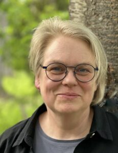 Catharina Coenen is facing the viewer, smiling, wearing a grey tshirt and black collared shirt in front of a tree/greenery background.
