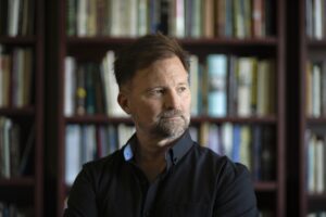 Christopher Bakken is looking to the viewers right posed in a black collared shirt in front of a blurred bookshelf.