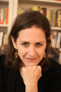Jessica Elisheva Emerson is facing the viewer, smiling, fist under chin. Emerson is wearing a black top and skull earings, posed in front of a bookshelf background.