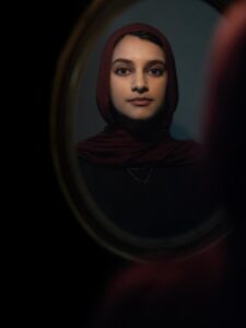 Viewer sees over Sarah Ghazal Ali's shoulder to reflection in an oval framed mirror. Ali is wearing a red head covering, black clothing, and necklace.