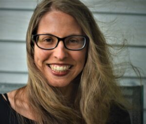 Jessie van Eerden is facing the viewer in front of a white siding background. Eerden is smiling, wearing a black top, black glasses, hair is being swept to the side by wind. 