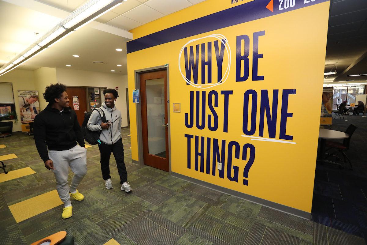 two students walk near a sign reading "Why be just one thing?"