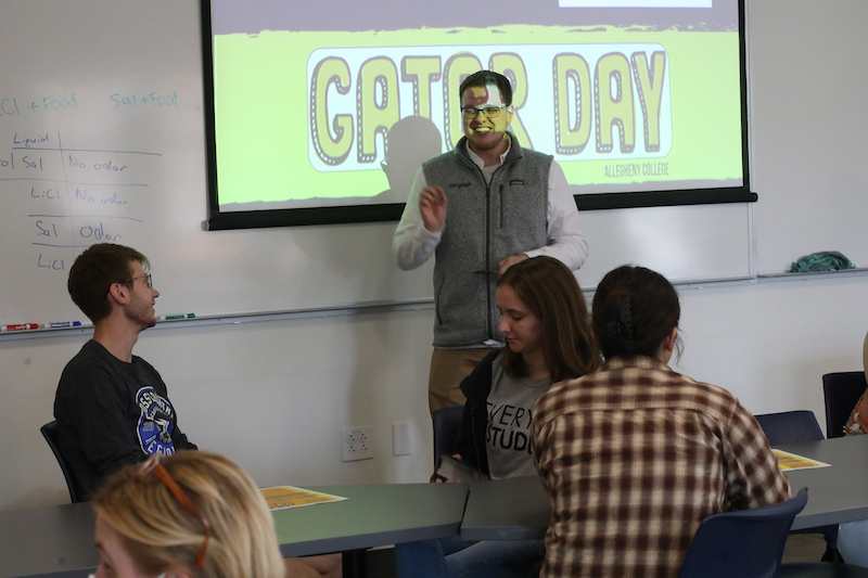 a person stands in front of a screen reading "Gator Day" with audience members looking on