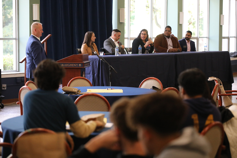 A speaker at a podium with five panelists at a table and audience members at tables in the foreground