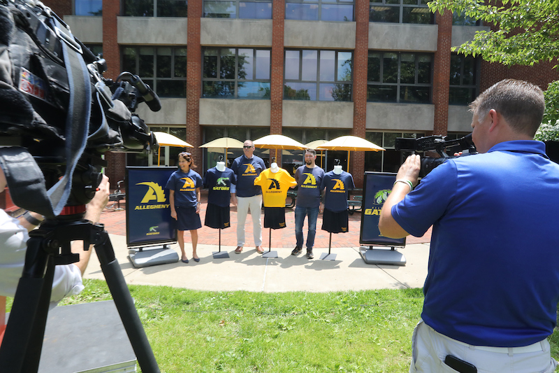 TV camera crews film three people standing with new T-shirts bearing the Allegheny College athletics logo