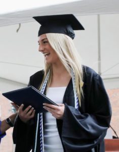 blonde female student smiling and wearning graduation robe and cap while holding diploma