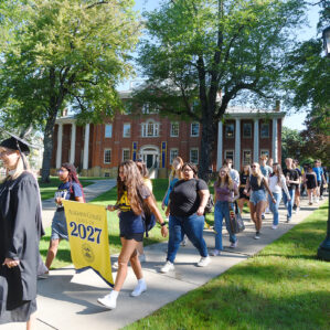 Students walking down the sidewalk in front of Bentley Hall, carrying a class flag and processing